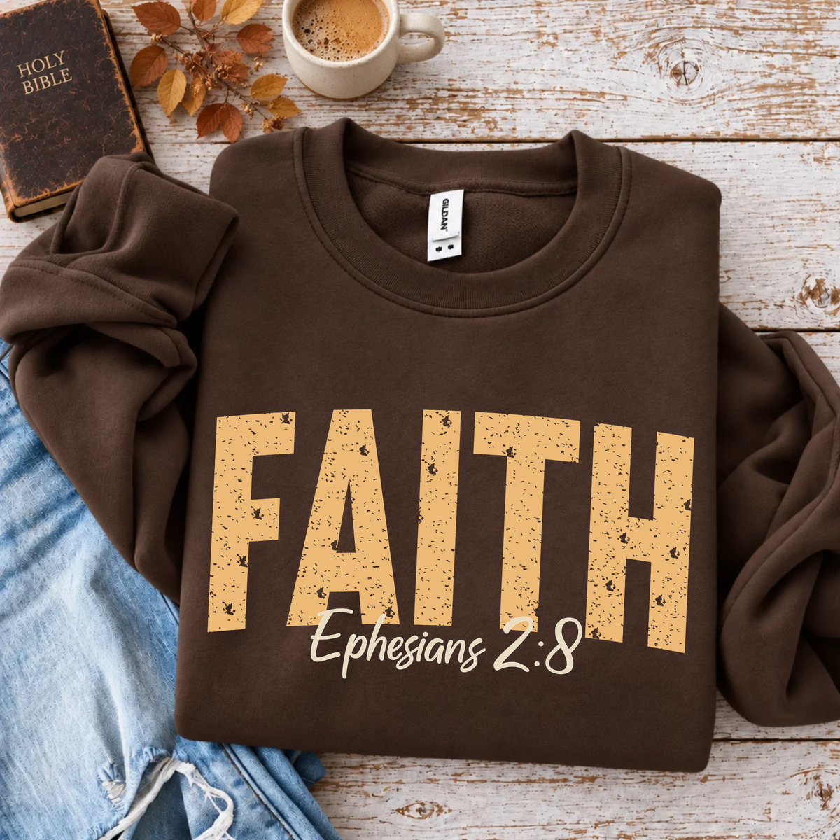 Brown crewneck sweatshirt flat lay with distressed gold FAITH lettering and white Ephesians 2:8 script, styled with a Holy Bible, coffee, autumn leaves, and blue jeans.