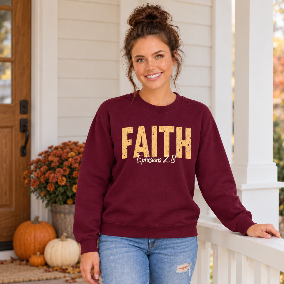 Woman wearing a maroon crewneck sweatshirt with distressed gold FAITH lettering and white Ephesians 2:8 script beside pumpkins and mums.