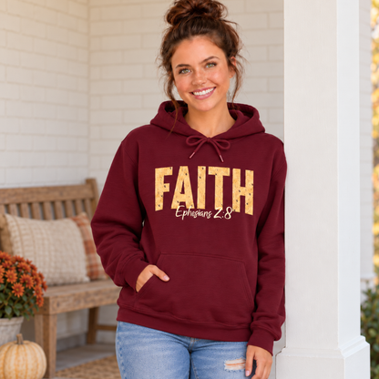 Woman modeling a maroon pullover hoodie featuring distressed gold FAITH text and white Ephesians 2:8 script on a cozy fall porch.