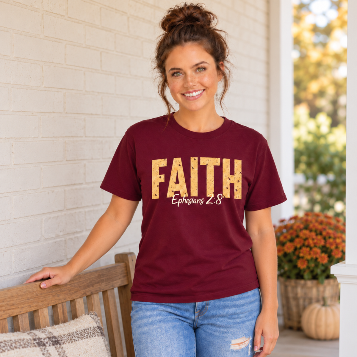 Woman wearing a maroon T-shirt with distressed gold FAITH lettering and white Ephesians 2:8 script near a porch bench, pumpkins, and mums.