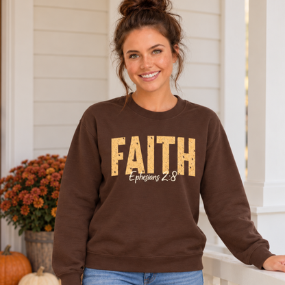 Woman wearing a brown crewneck sweatshirt with distressed gold FAITH lettering and white Ephesians 2:8 script on a fall porch with pumpkins and mums.