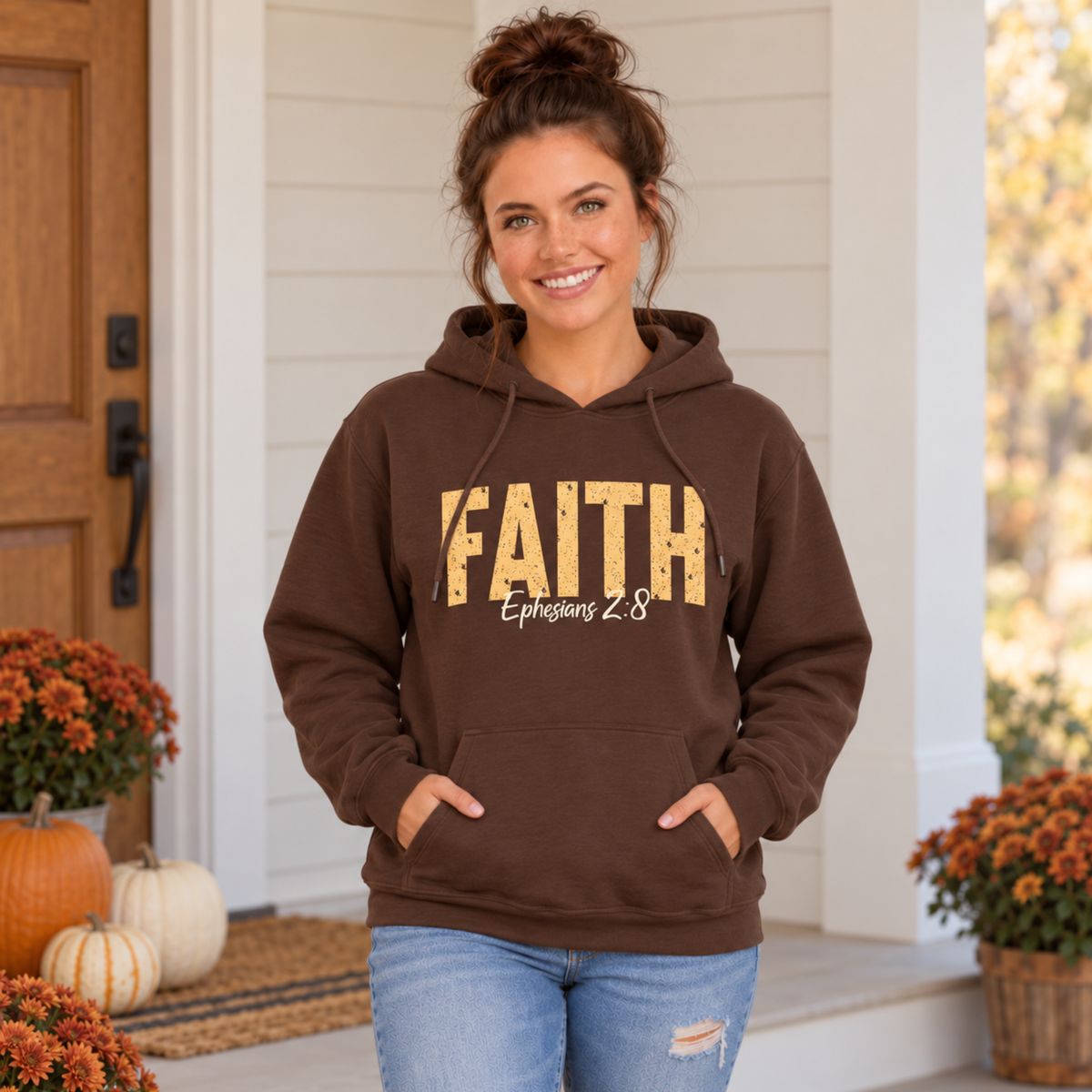 Woman modeling a brown pullover hoodie featuring distressed gold FAITH text and white Ephesians 2:8 script beside pumpkins and orange fall flowers.