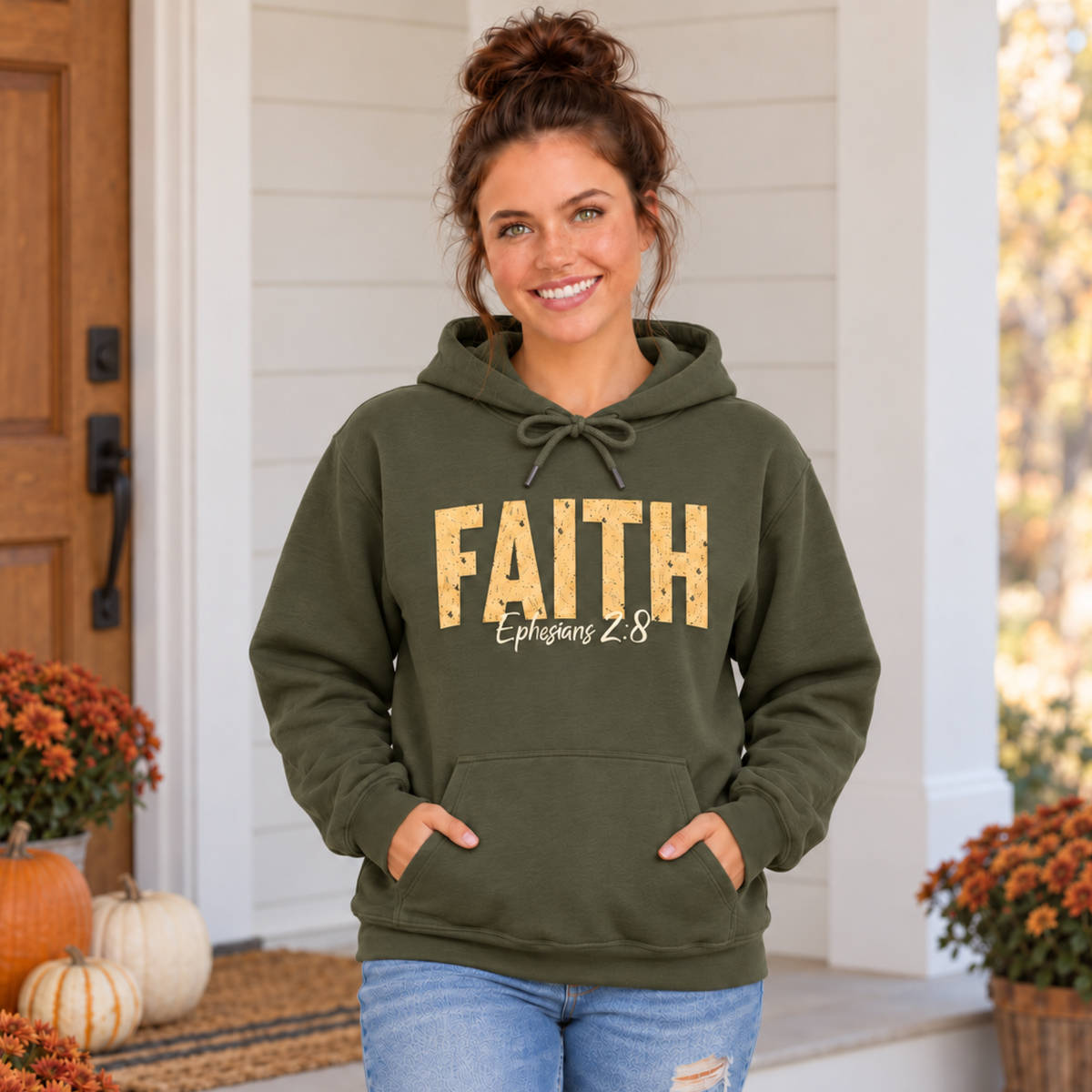 Woman wearing a military green pullover hoodie with distressed gold FAITH lettering and white Ephesians 2:8 script on a decorated fall porch.