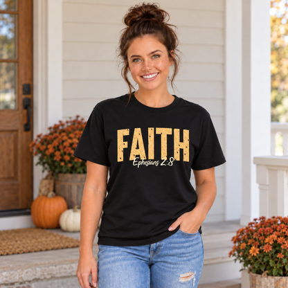 Woman modeling a black T-shirt with distressed gold FAITH text and white Ephesians 2:8 script in front of pumpkins and orange mums.