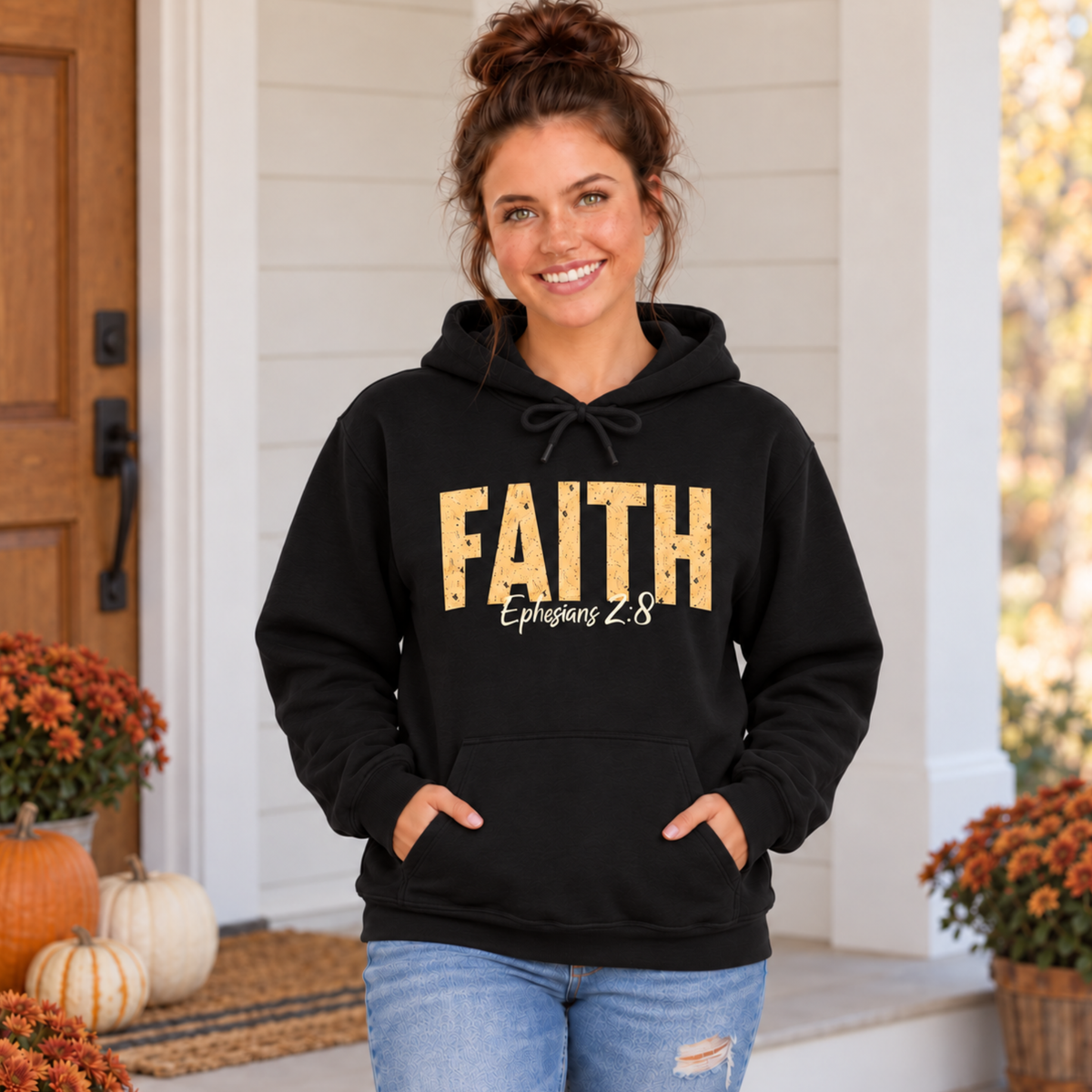 Woman modeling a black pullover hoodie with distressed gold FAITH text and white Ephesians 2:8 script among fall pumpkins and flowers.