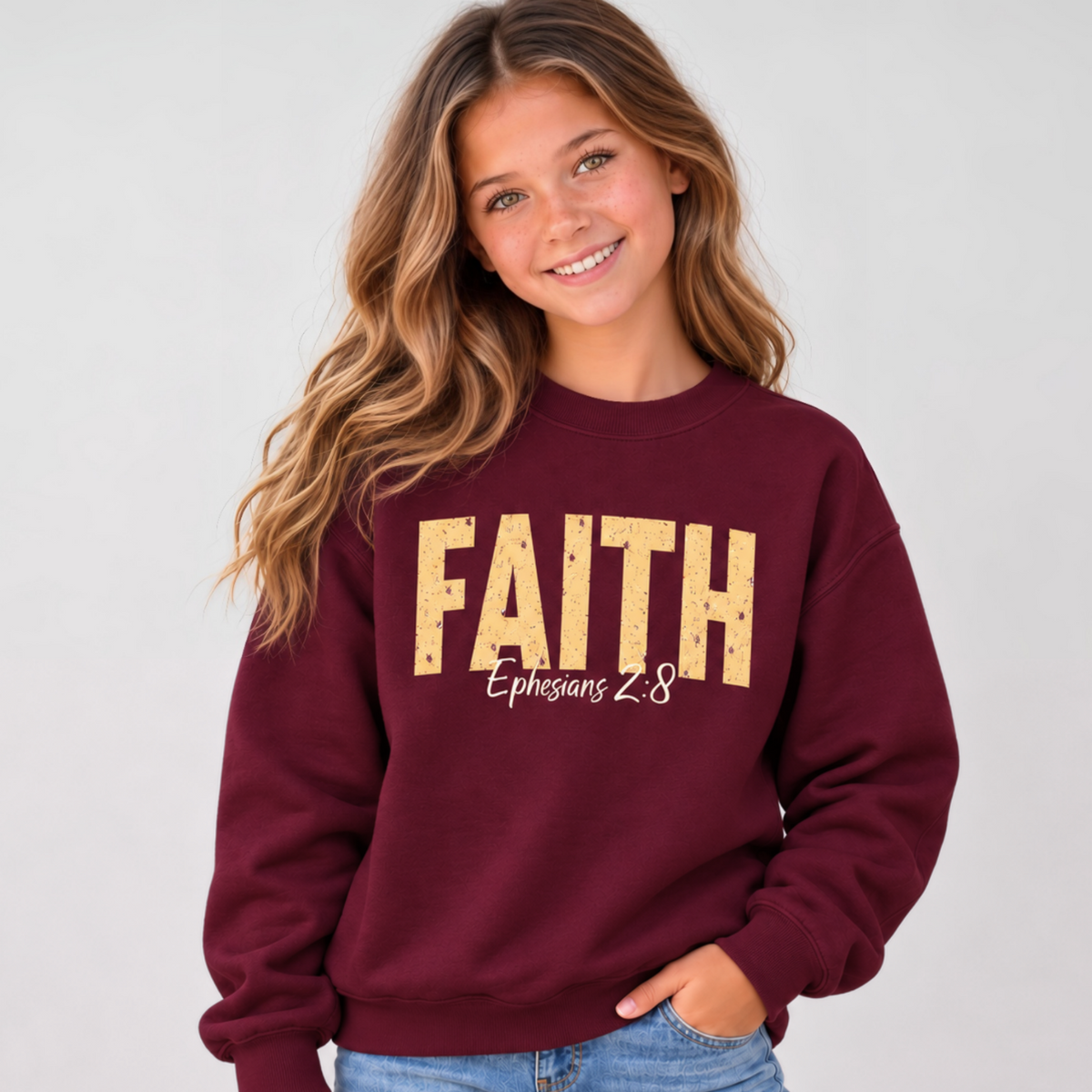 Young model wearing a maroon youth crewneck sweatshirt with distressed gold FAITH lettering and white Ephesians 2:8 script against a simple light gray background.