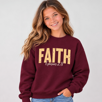 Young model wearing a maroon youth crewneck sweatshirt with distressed gold FAITH lettering and white Ephesians 2:8 script against a simple light gray background.