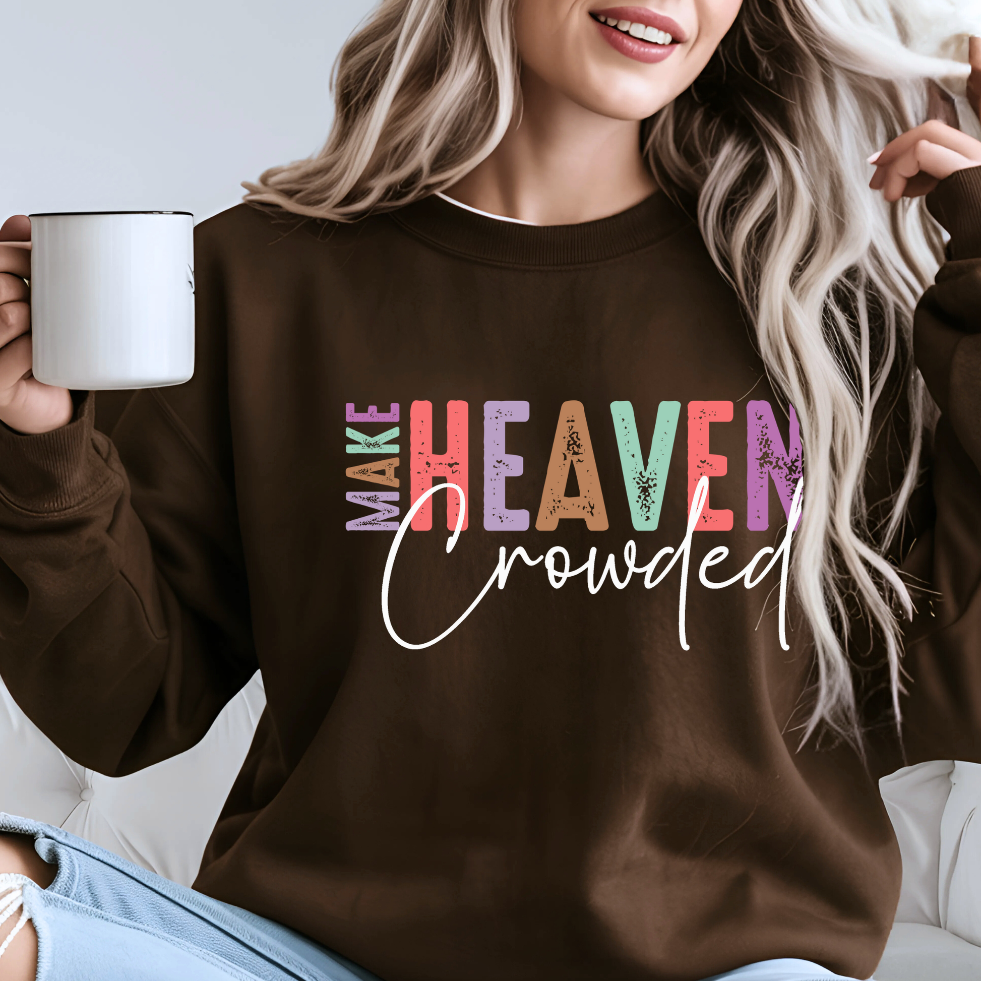 Woman wearing brown Christian sweatshirt with bold multicolor “Make Heaven Crowded” design, holding coffee mug. Religious inspirational faith crewneck for women.