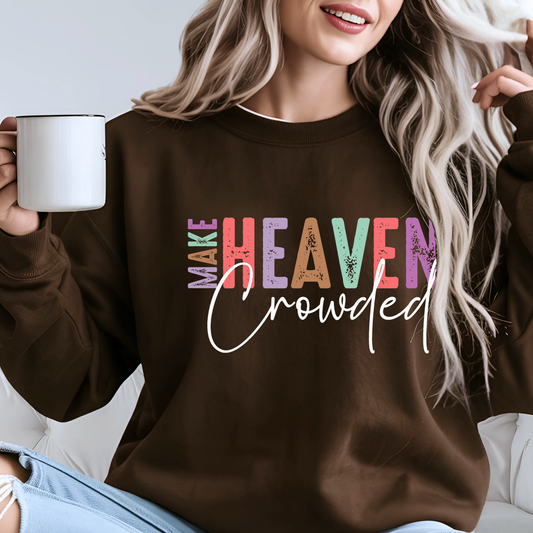 Woman wearing brown Christian sweatshirt with bold multicolor “Make Heaven Crowded” design, holding coffee mug. Religious inspirational faith crewneck for women.
