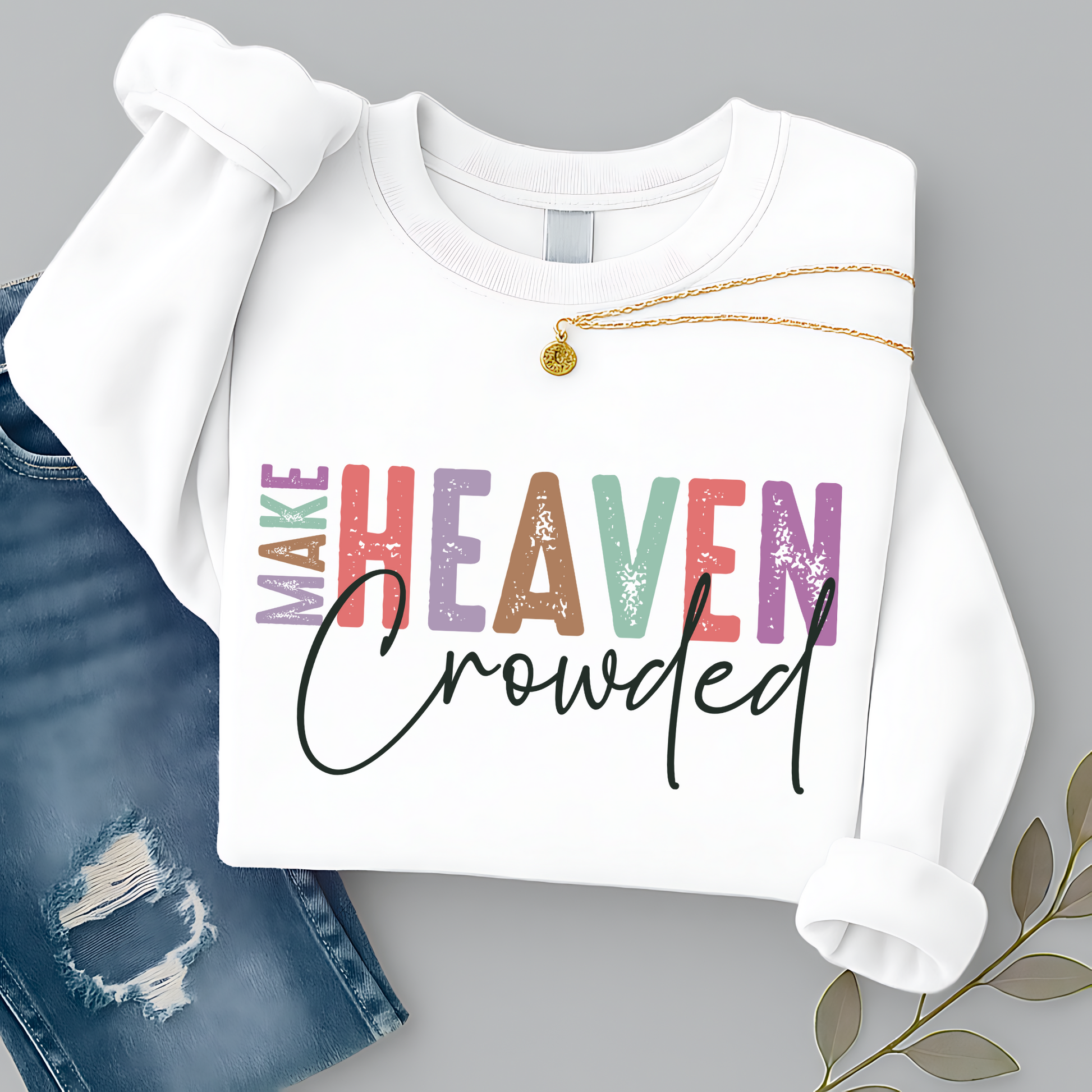White unisex sweatshirt with colorful “Make Heaven Crowded” Christian graphic design, styled with necklace and jeans. Faith sweatshirt for women and men, inspirational religious apparel.