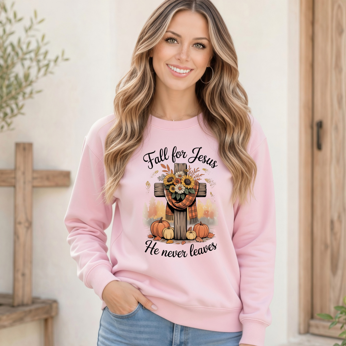 Woman wearing a light pink crewneck sweatshirt with the Fall for Jesus He Never Leaves cross, pumpkin, and sunflower design in a rustic indoor setting.