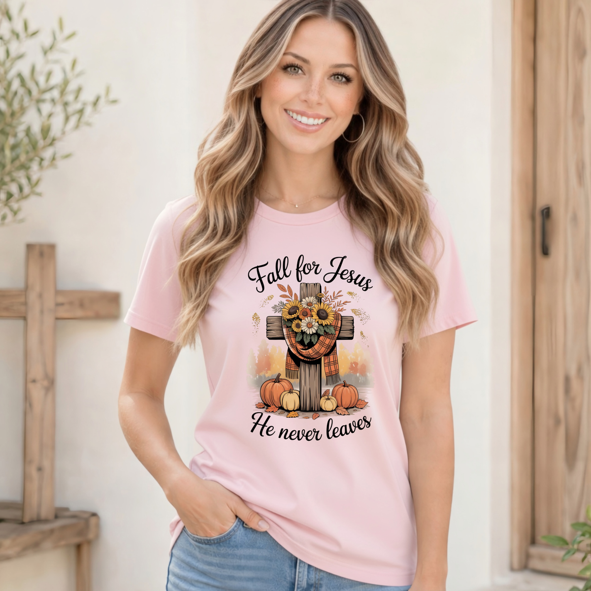 Smiling woman wearing a light pink T-shirt with the Fall for Jesus He Never Leaves cross, pumpkin, and sunflower design near a rustic wooden doorway.