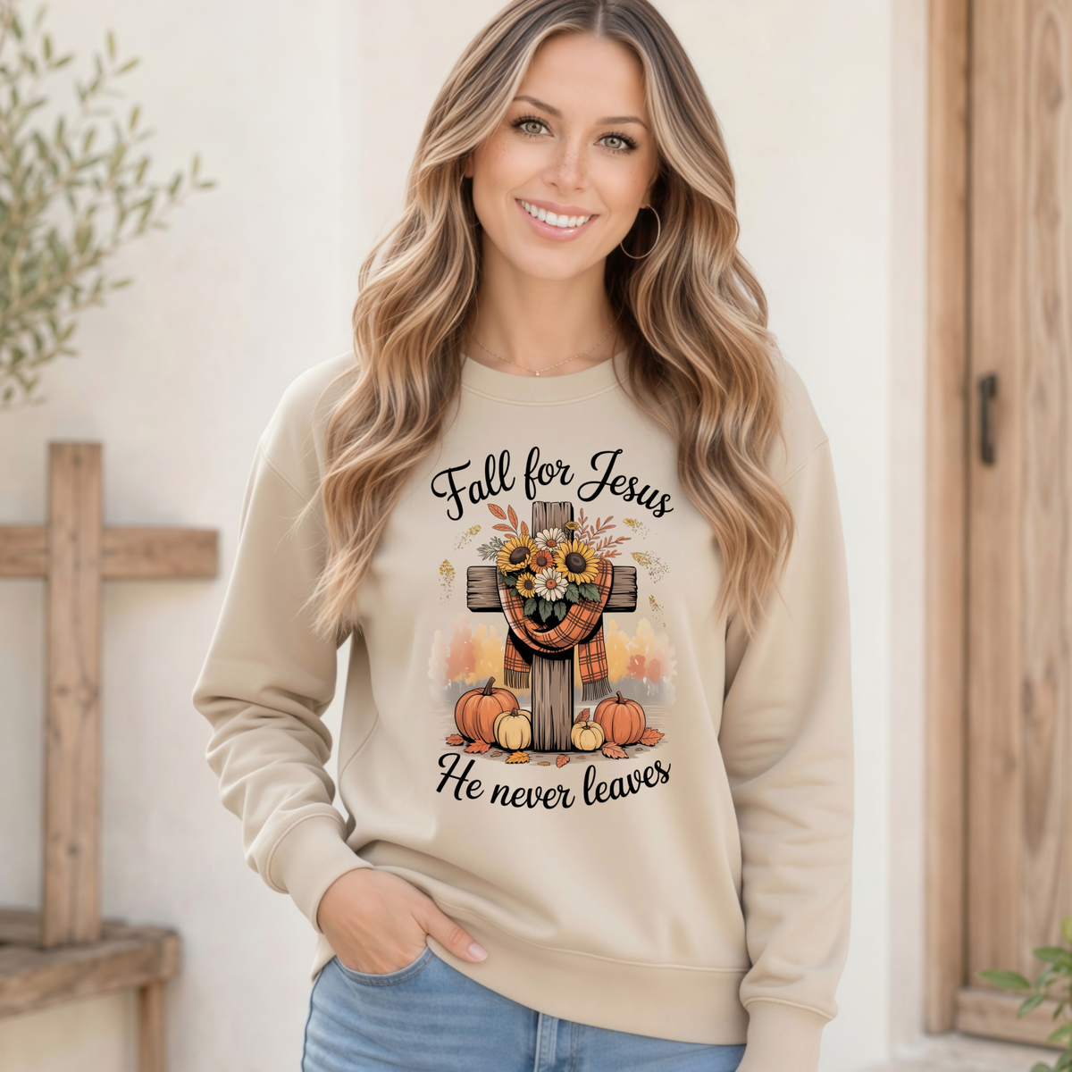 Woman wearing a Sand crewneck sweatshirt with the Fall for Jesus He Never Leaves design featuring a cross, pumpkins, and sunflowers by a rustic wooden doorway.