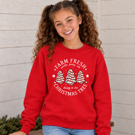 Girl modeling a red Farm Fresh Christmas Tree Cakes sweatshirt with three frosted tree graphics against a light wall.