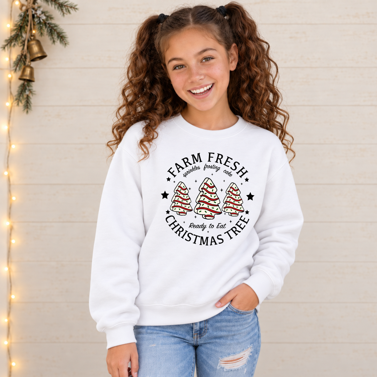 Girl modeling a white Farm Fresh Christmas Tree Cakes sweatshirt against a light holiday wall.