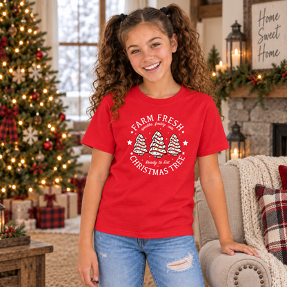 Girl modeling a red Farm Fresh Christmas Tree Cakes T-shirt in front of a Christmas tree.