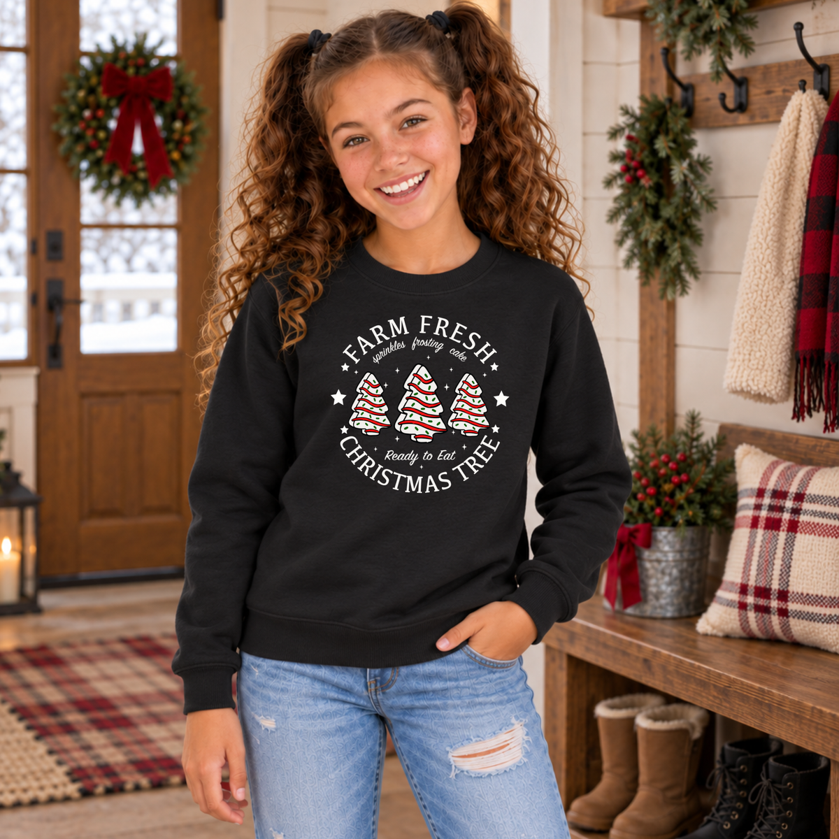 Girl wearing a black Farm Fresh Christmas Tree Cakes sweatshirt in a cozy holiday room.