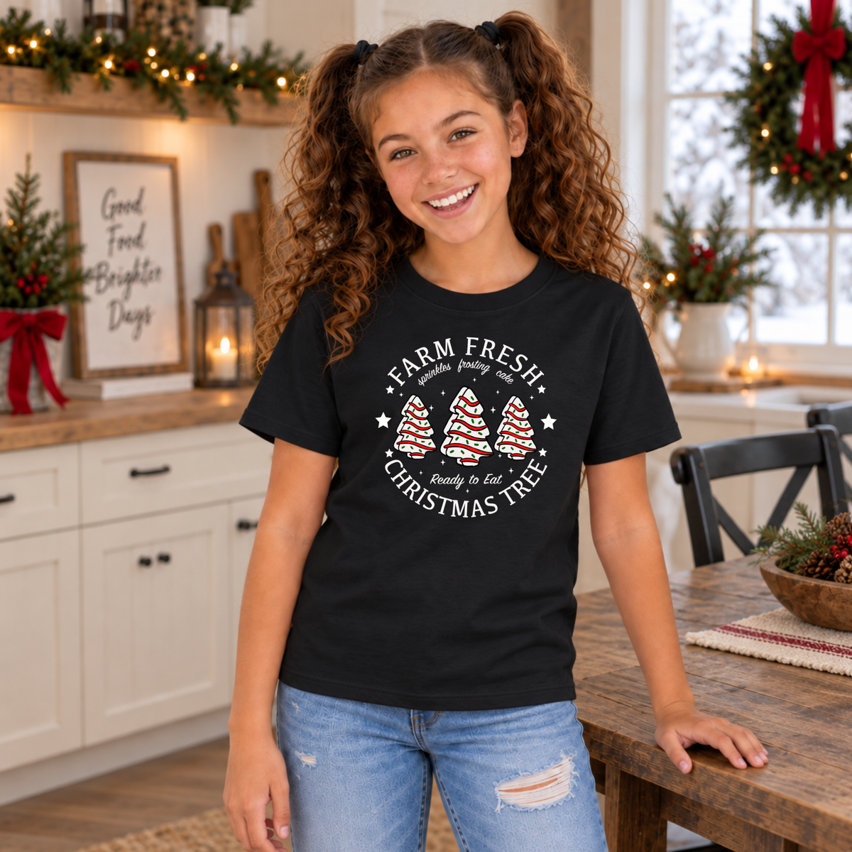 Girl wearing a black Farm Fresh Christmas Tree Cakes T-shirt beside a holiday dining table.