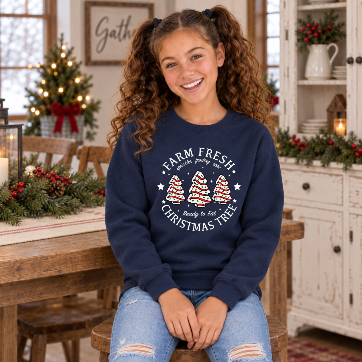 Girl seated in a navy Farm Fresh Christmas Tree Cakes sweatshirt by a decorated farmhouse table.
