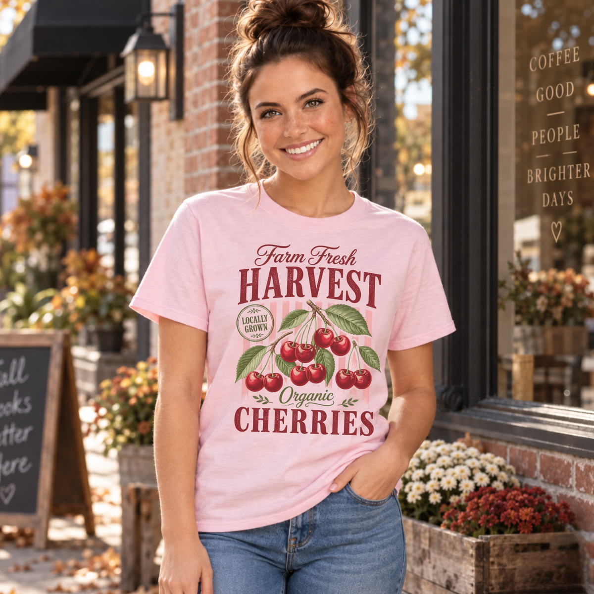 Woman in a light pink Farm Fresh Harvest Organic Cherries T-shirt outside a coffee shop with flower planters.