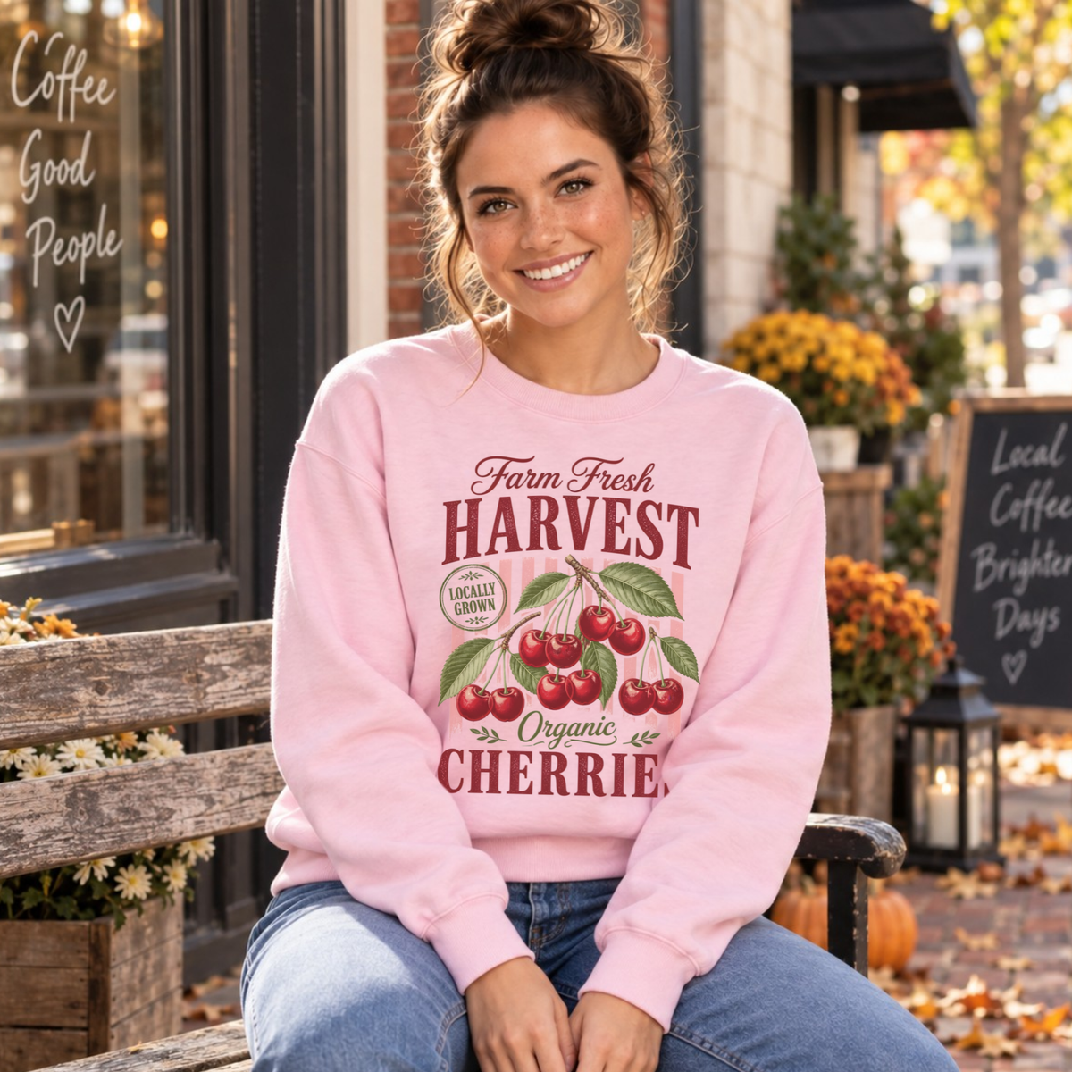 Woman seated on a bench outside a coffee shop wearing a light pink Farm Fresh Harvest Organic Cherries sweatshirt.