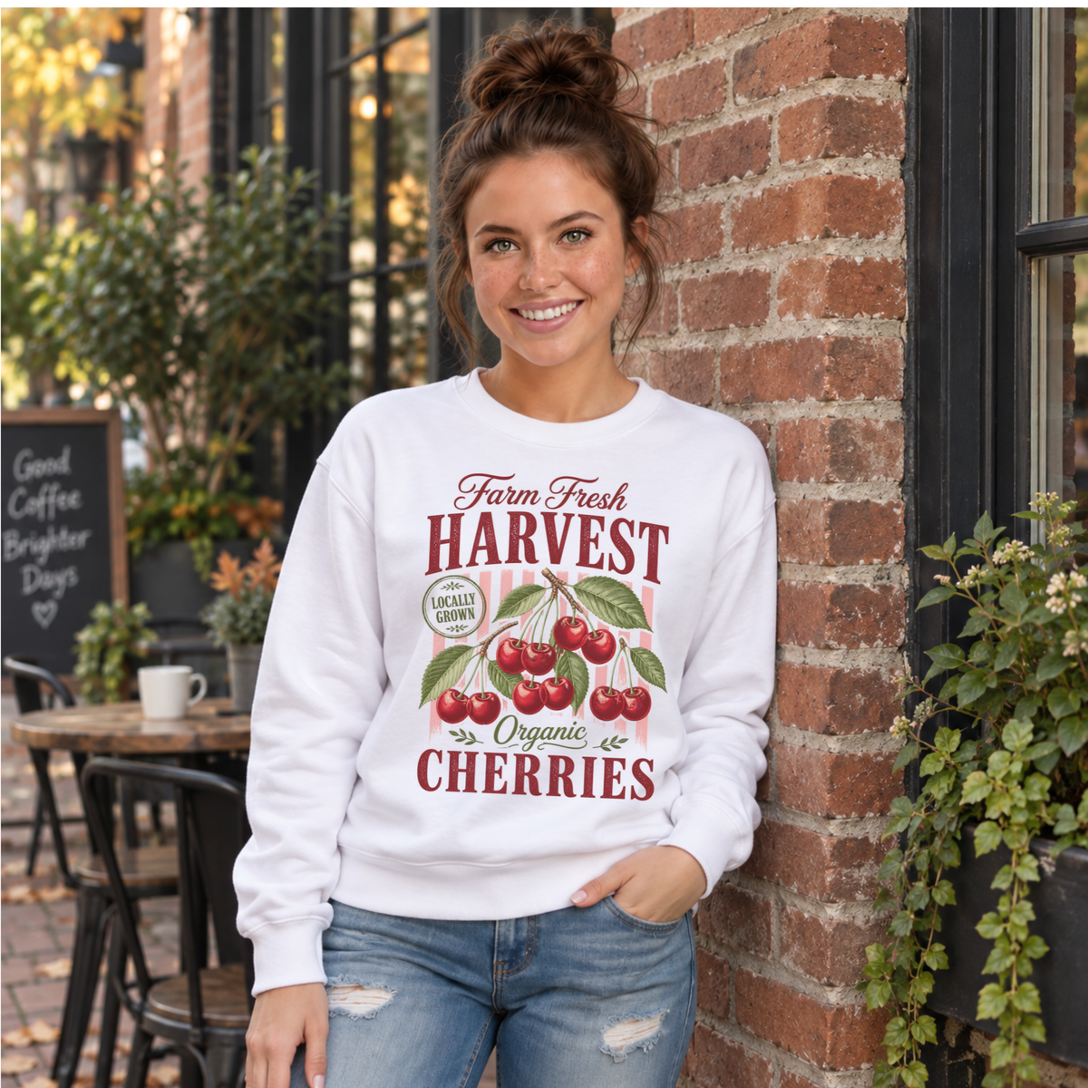 Woman in a white Farm Fresh Harvest Organic Cherries sweatshirt leaning against a brick coffee shop wall.