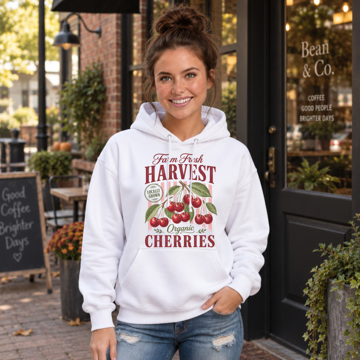 Woman wearing a white Farm Fresh Harvest Organic Cherries hoodie outside a brick coffee shop.
