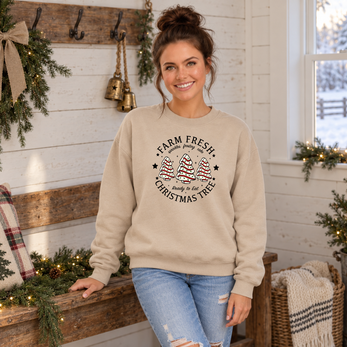 Woman modeling a sand Farm Fresh Christmas Tree Cakes sweatshirt beside a decorated farmhouse bench.