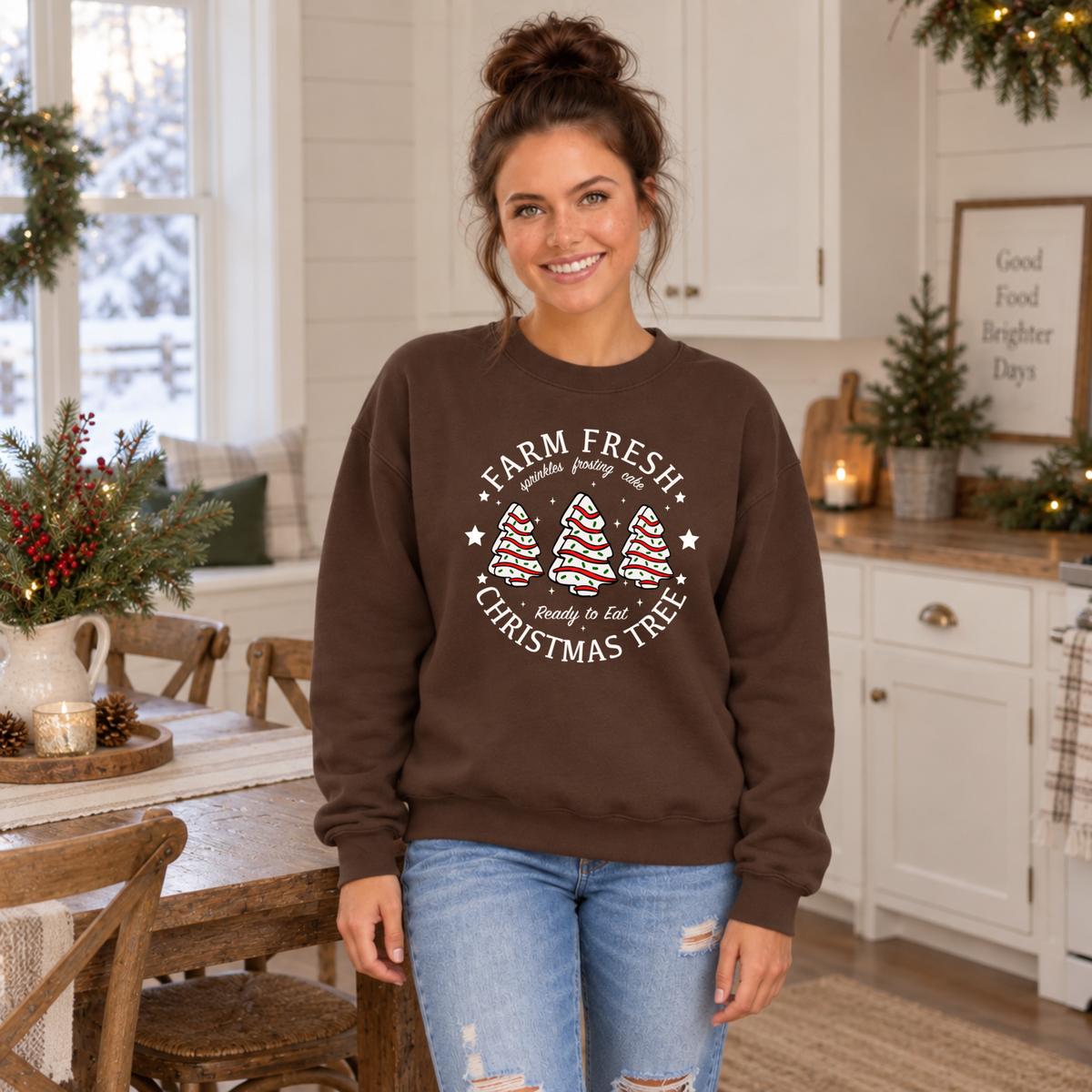 Woman modeling a brown Farm Fresh Christmas Tree Cakes sweatshirt in a festive farmhouse kitchen.