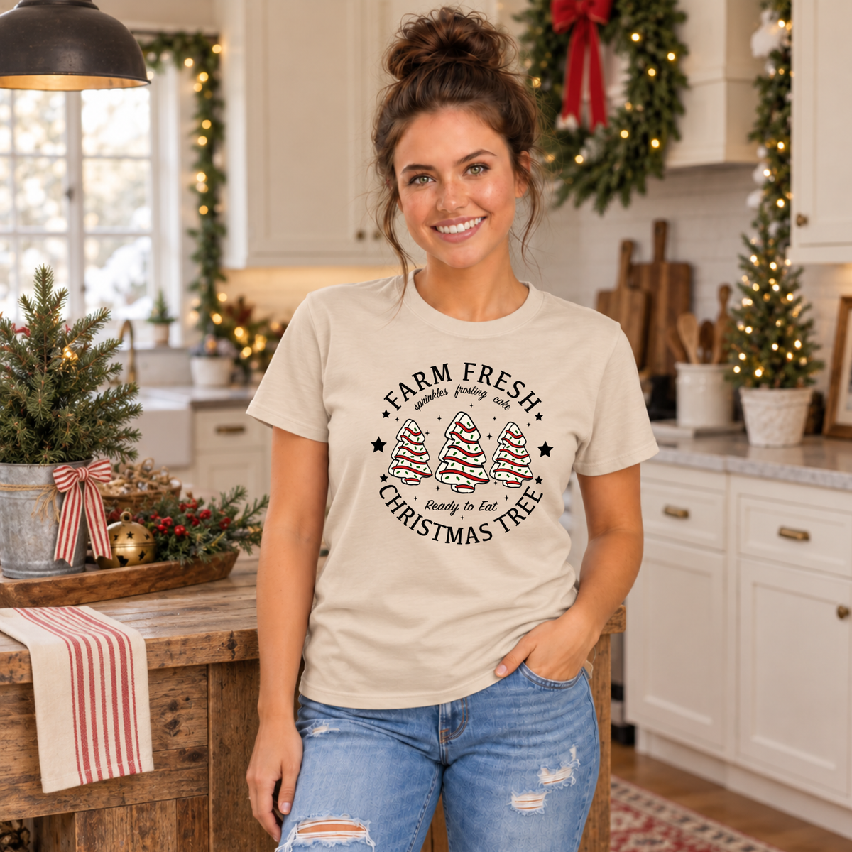 Woman wearing a sand Farm Fresh Christmas Tree Cakes T-shirt in a festive kitchen.