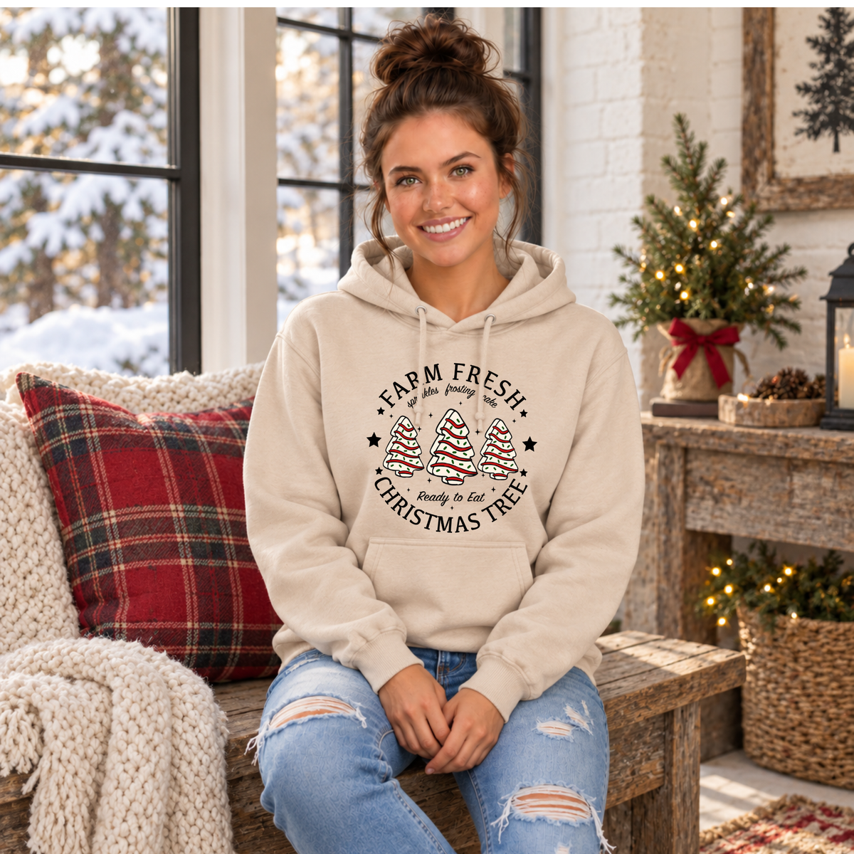 Woman seated in a sand Farm Fresh Christmas Tree Cakes hoodie beside holiday decor.