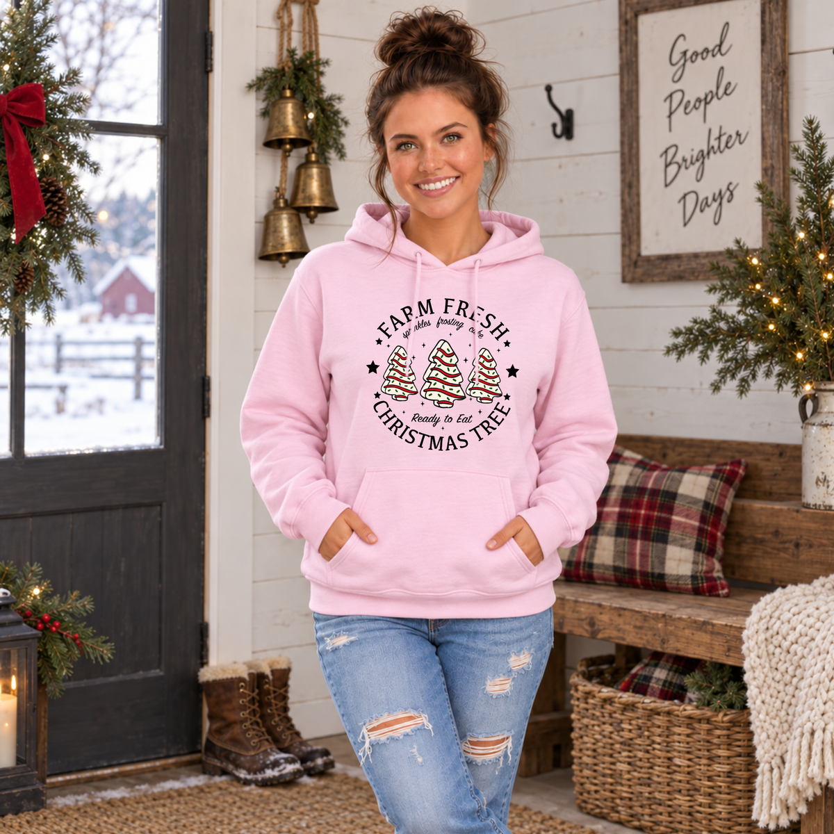 Woman wearing a light pink Farm Fresh Christmas Tree Cakes hoodie in a festive farmhouse room.