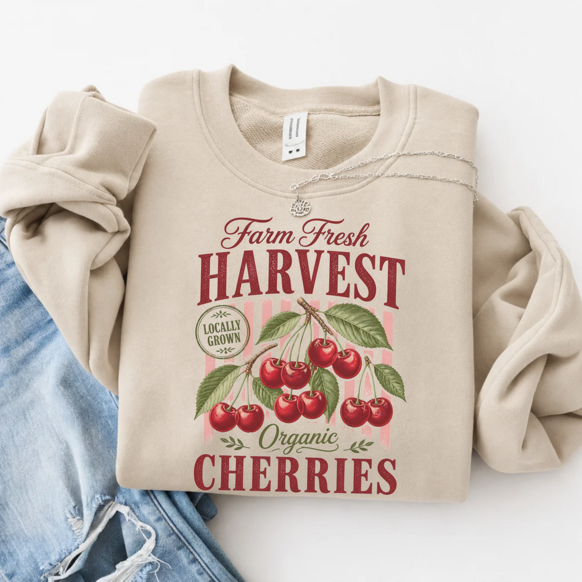 Sand crewneck sweatshirt flat lay with a vintage Farm Fresh Harvest Organic Cherries graphic, styled with blue denim on a white background.