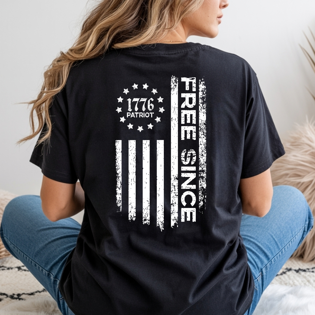 Seated woman shown from behind in a black T-shirt with a large distressed white Free Since flag graphic and 1776 Patriot emblem