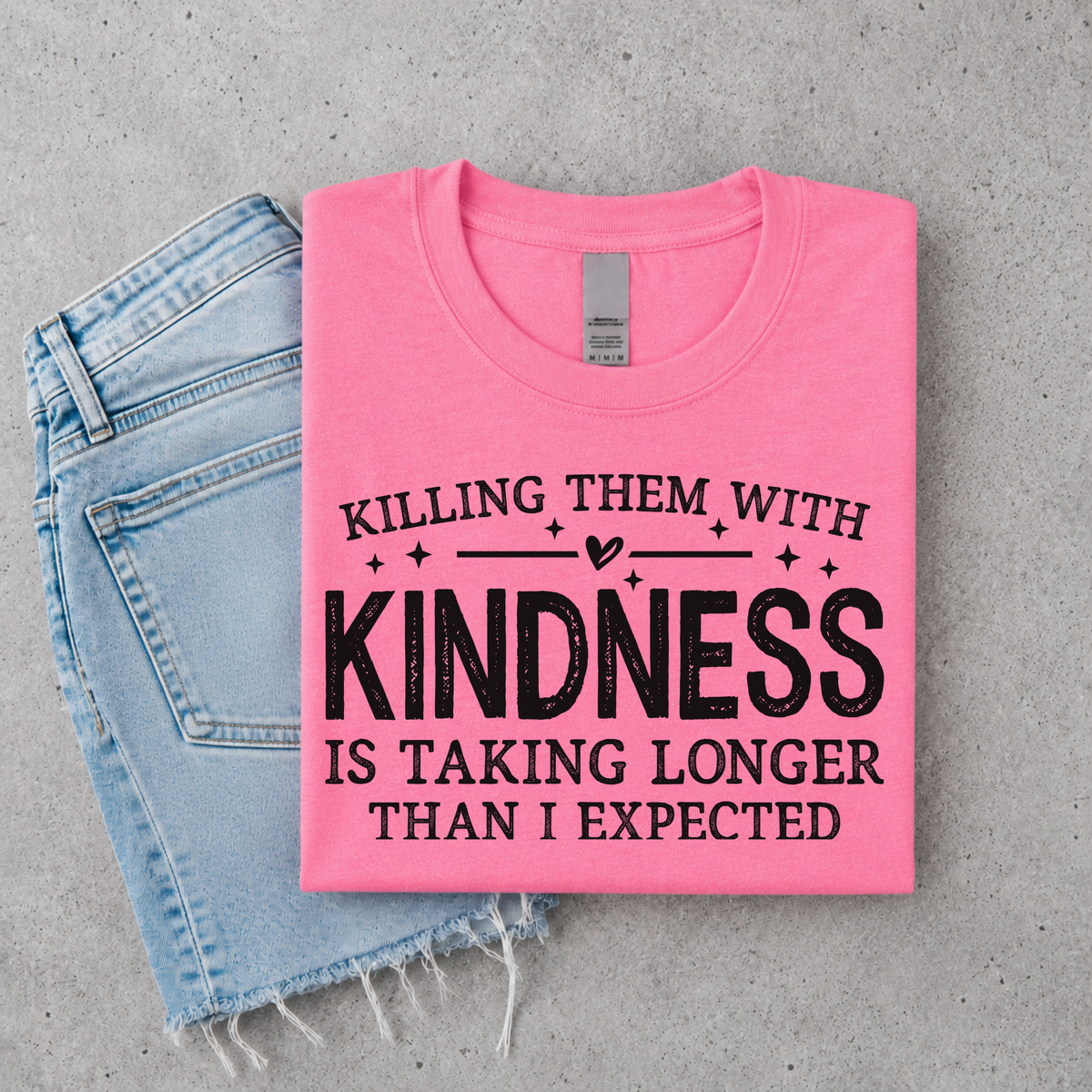 Bright Pink Killing Them With Kindness T-shirt flat lay with rolled sleeves, and distressed denim shorts on a gray background.