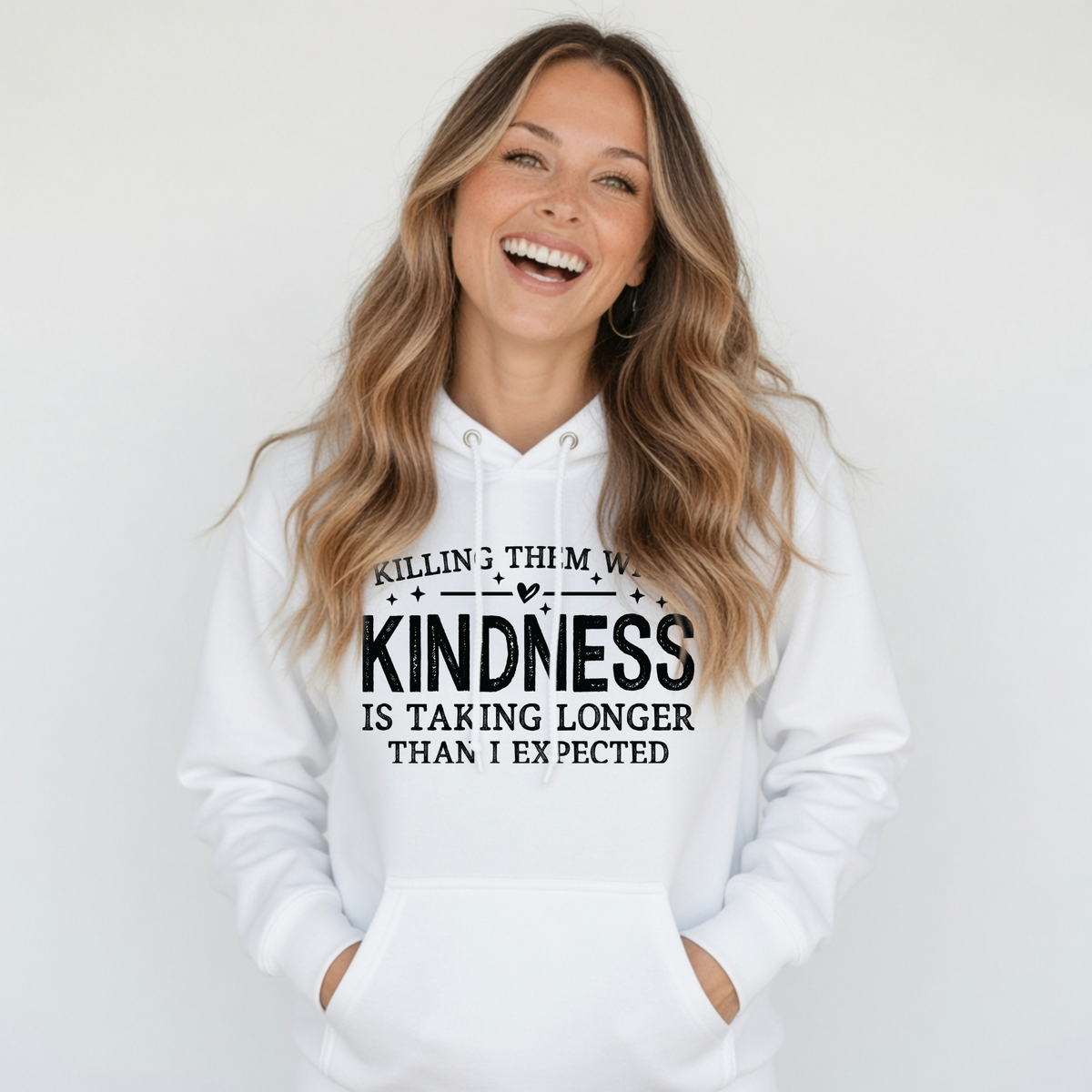 Laughing woman wearing a white Killing Them With Kindness hoodie against a light neutral background.