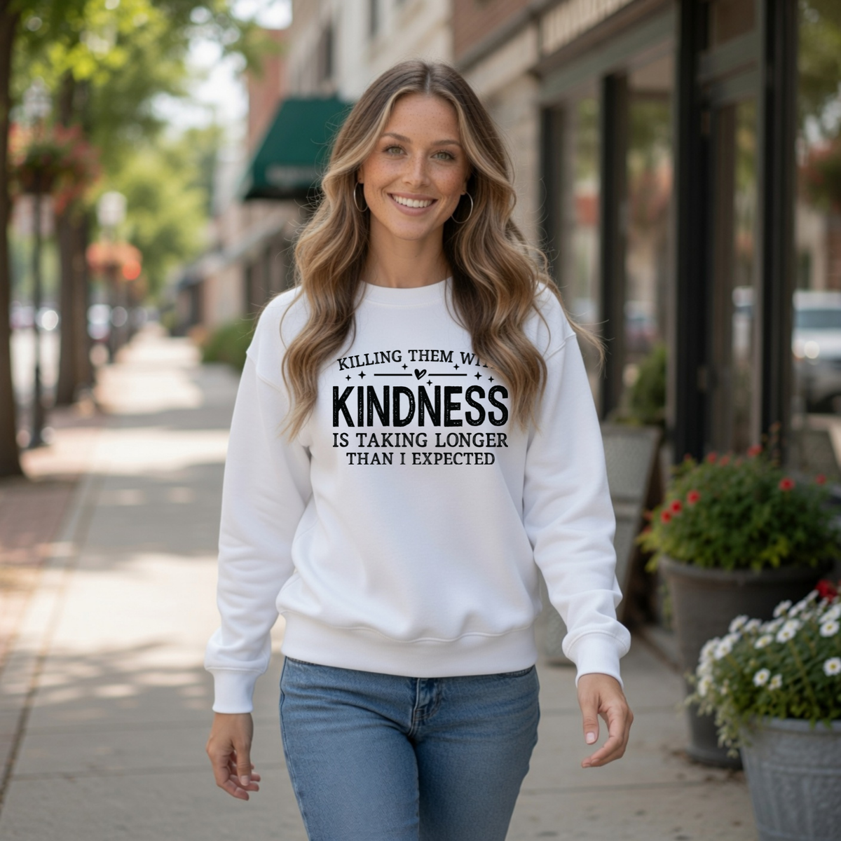 Woman walking outdoors in a white Killing Them With Kindness crewneck sweatshirt with jeans on a small-town sidewalk.