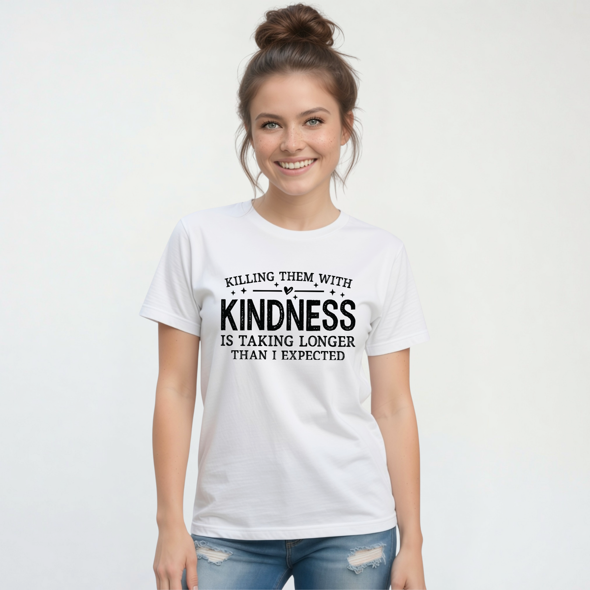 Smiling woman modeling a white Killing Them With Kindness T-shirt with distressed jeans against a white background.