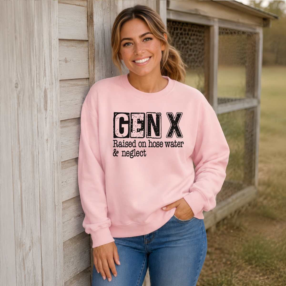 Woman leaning against a farmhouse wall in a light pink GEN X Raised on hose water & neglect sweatshirt.