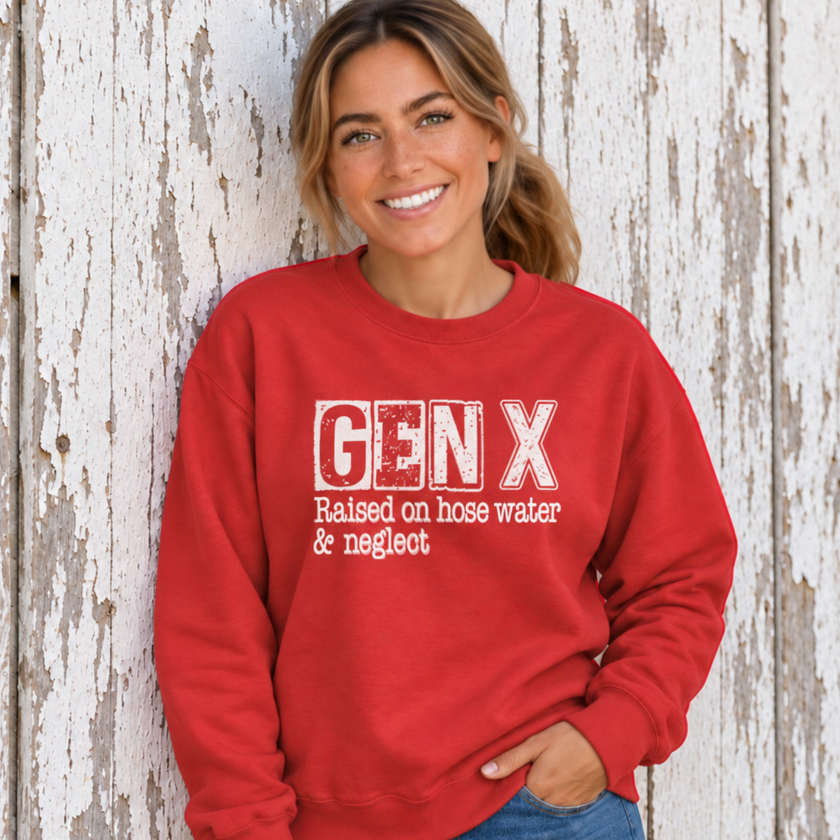 Woman wearing a red GEN X Raised on hose water & neglect sweatshirt against a distressed white barn wall.