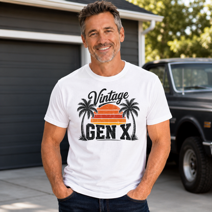 Man in a white Vintage Gen X T-shirt with palm tree sunset design standing near a garage and vintage truck.