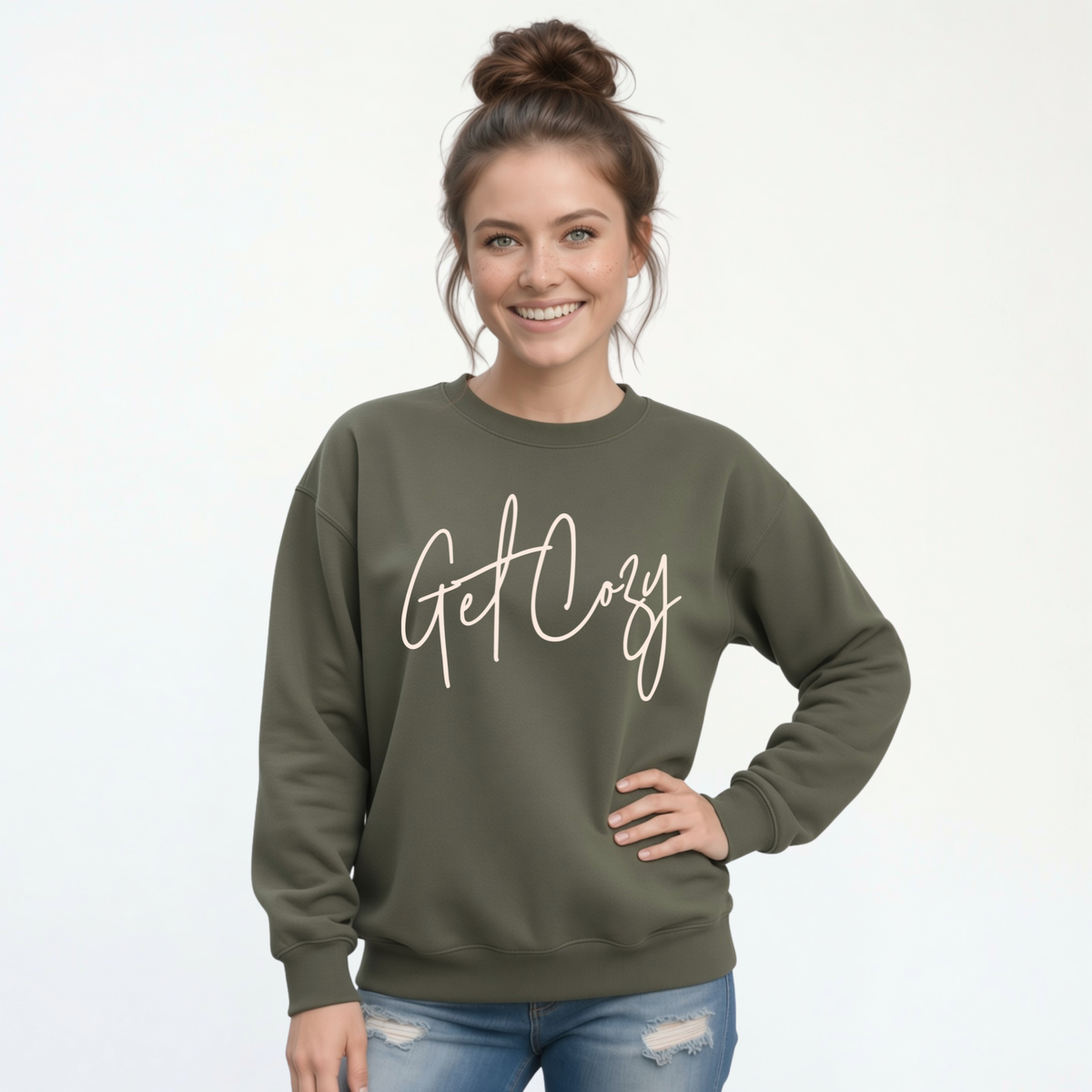 Woman wearing a military green crewneck sweatshirt with a minimalist Get Cozy script design, paired with distressed blue jeans on a white background.