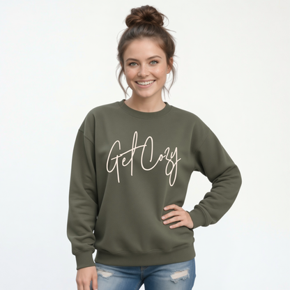 Woman wearing a military green crewneck sweatshirt with a minimalist Get Cozy script design, paired with distressed blue jeans on a white background.