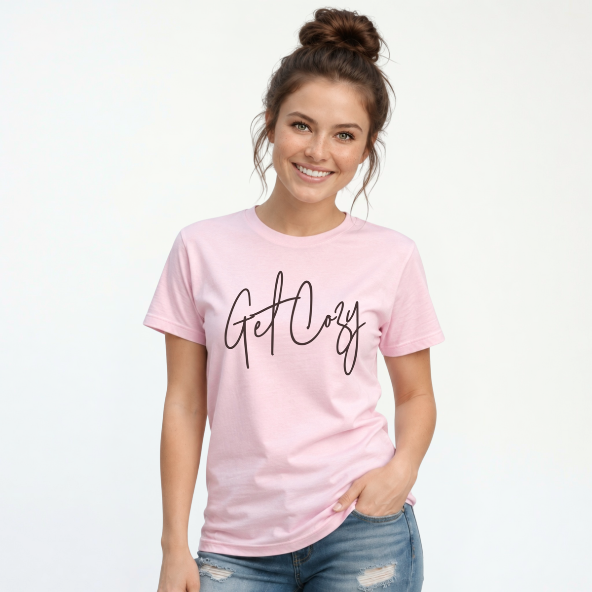 Woman wearing a light pink T-shirt with a minimalist Get Cozy script design, paired with distressed blue jeans on a white background.