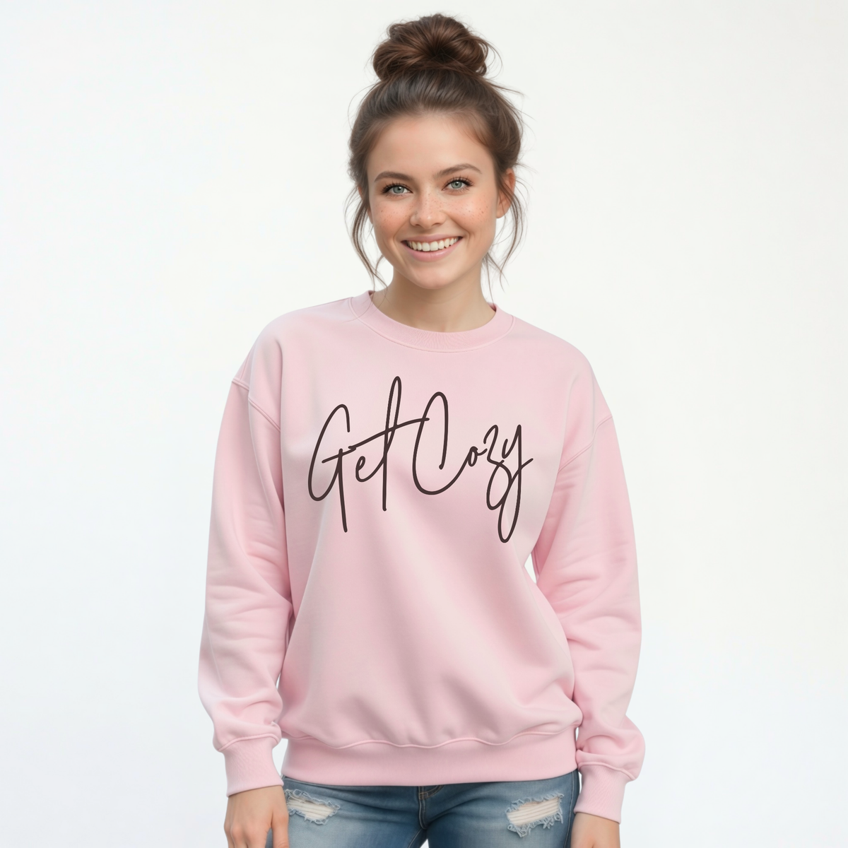 Woman wearing a light pink crewneck sweatshirt with a minimalist Get Cozy script design, paired with distressed blue jeans on a white background.