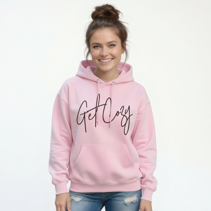 Woman wearing a light pink pullover hoodie with a minimalist Get Cozy script design, paired with blue jeans on a white background.
