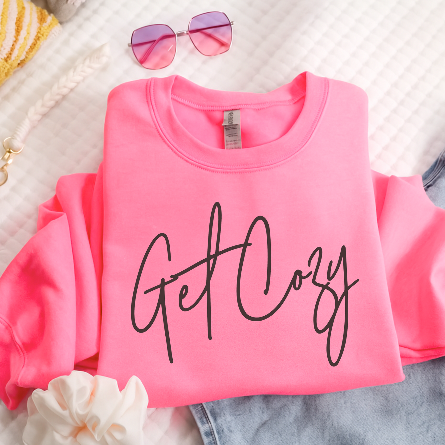 Pink sweatshirt with 'Get Cozy' text on a textured surface with sunglasses and flowers.