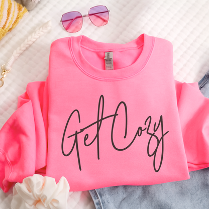 Pink sweatshirt with 'Get Cozy' text on a textured surface with sunglasses and flowers.