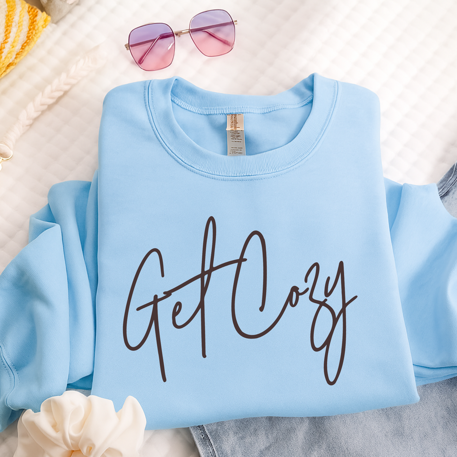 Light blue sweatshirt with 'Get Cozy' text on a white surface with sunglasses and flowers.