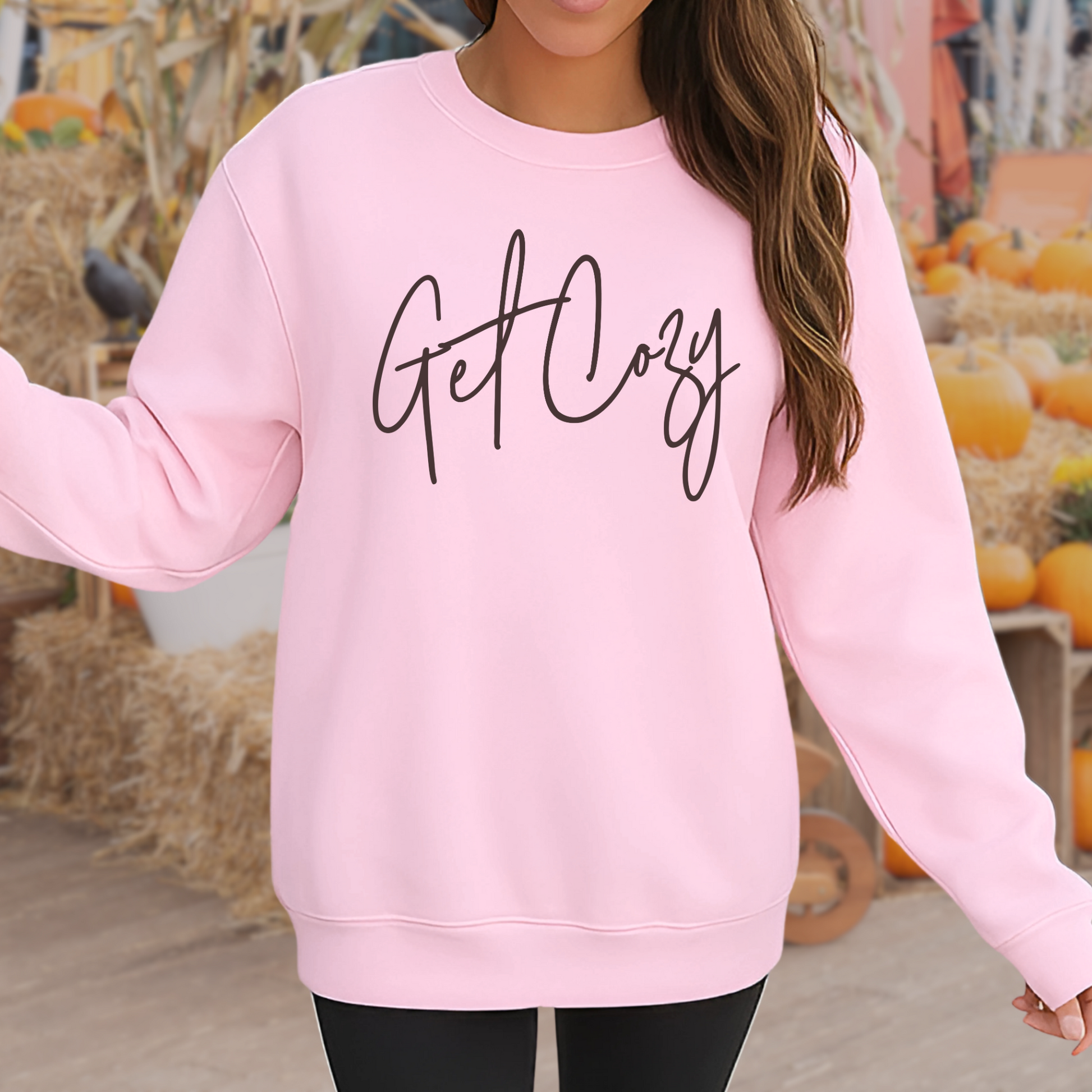 Person wearing a pink sweatshirt with 'Get Cozy' text in an autumn setting with pumpkins and hay.